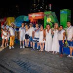 Un grupo de personas posan juntas por la noche delante de unas coloridas y grandes letras "Cartagena". Están vestidos con ropa blanca y de colores claros, sonríen y están de pie sobre una cubierta de madera con las luces de la ciudad de fondo.