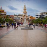 La gente pasea y monta en bicicleta por una amplia plaza con una estatua central y una torre del reloj, rodeada de árboles y edificios de colores bajo un cielo nublado.