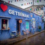 Un edificio azul vibrante con sillas de colores, banderas y arte mural muestra las palabras I ♥ Getsemaní en letras grandes en su fachada, a lo largo de una calle húmeda en Cartagena, Colombia.