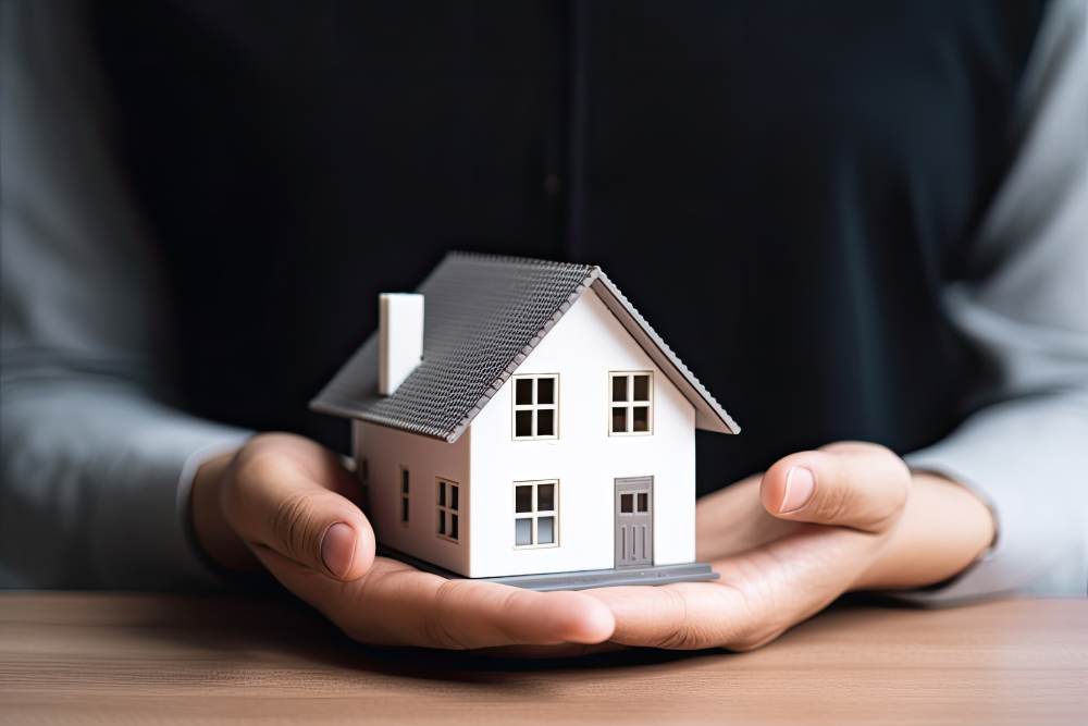 Una persona vestida de blanco y negro sostiene suavemente una pequeña maqueta de casa, símbolo de protección y vivienda propia o de conceptos inmobiliarios como Crédito Comfamiliar, con una superficie de madera en primer plano.