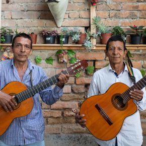 Dos hombres sostienen sendas guitarras acústicas frente a un muro de ladrillo decorado con pequeñas macetas y vegetación, combinando su estilo con el encanto de 14 estaciones Balboa. Ambos llevan camisas abotonadas y miran a la cámara.