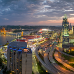 Vista aérea del centro de Cincinnati, Estados Unidos, al atardecer, mostrando edificios iluminados, autopistas y el río Ohio con puentes al fondo bajo un cielo nublado.