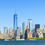 La imagen muestra el horizonte de Nueva York en Estados Unidos con altos rascacielos, incluido el One World Trade Center, bajo un cielo azul despejado, visto desde el otro lado del río con barcos en el agua.