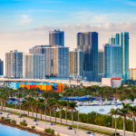 Paisaje urbano de Miami en los Estados Unidos con altos edificios modernos, palmeras a lo largo del paseo marítimo, un puerto deportivo lleno de barcos y un puente que cruza el agua bajo un cielo parcialmente nublado.