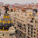 Vista aérea del edificio Metrópolis, con su cúpula negra ornamentada y su estatua alada, rodeado de edificios históricos y calles del centro de Madrid, España.