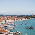 Vista aérea del paseo marítimo de Venecia con edificios históricos, calles abarrotadas y numerosos barcos y transbordadores que navegan por el Gran Canal azul verdoso bajo un cielo despejado.