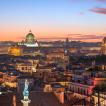 Vista de Roma al atardecer, con los tejados de la ciudad en primer plano y las cúpulas iluminadas de las iglesias históricas, incluida la Basílica de San Pedro, visibles al fondo contra un cielo colorido.