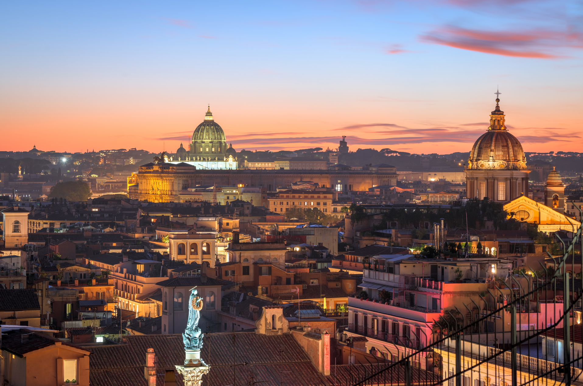 Vista de Roma al atardecer, con los tejados de la ciudad en primer plano y las cúpulas iluminadas de las iglesias históricas, incluida la Basílica de San Pedro, visibles al fondo contra un cielo colorido.