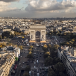 Vista aérea del Arco del Triunfo de París, rodeado de tráfico y edificios históricos, con un espectacular cielo nublado.