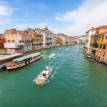 Barcos recorren un canal flanqueado por edificios de colores en Venecia, Italia, bajo un cielo parcialmente nublado. La gente espera en las paradas de los autobuses acuáticos a lo largo del paseo marítimo. El agua refleja el animado paisaje urbano.