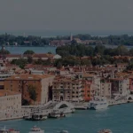 Una vista panorámica de Venecia con edificios de tejados rojos a lo largo del paseo marítimo, varios barcos en el agua azul verdosa y árboles verdes al fondo bajo un cielo parcialmente nublado.