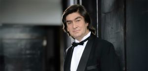 Un hombre de pelo oscuro de media melena, vestido con esmoquin negro con pajarita y camisa blanca, permanece de pie en el interior apoyado en una pared oscura, mirando con confianza a la cámara: una imagen que recuerda a Fausto en una Consotá Serenata.