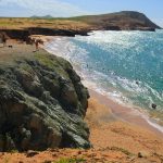 Unos acantilados rocosos dan a una playa de arena con olas turquesas. La gente está de pie en los acantilados y nadando en el océano bajo un cielo claro y soleado. La costa se extiende en la distancia con una vegetación verde.