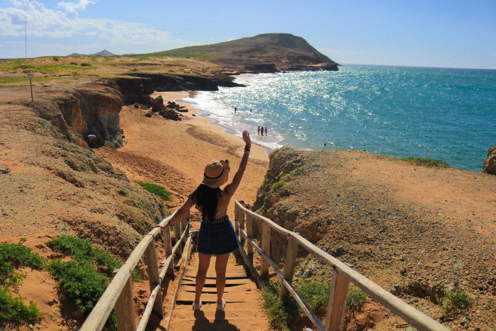 Una mujer con sombrero baja unas escaleras de madera hacia una playa de arena con agua turquesa, acantilados rocosos y algunas personas a lo lejos bajo un cielo claro y brillante.