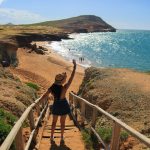 Una mujer con sombrero baja unas escaleras de madera hacia una playa de arena con agua turquesa, acantilados rocosos y algunas personas a lo lejos bajo un cielo claro y brillante.