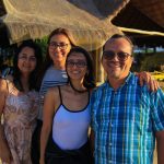 Cuatro adultos permanecen juntos, sonriendo al aire libre bajo la luz del sol. Están delante de un edificio rústico con tejado de paja. Al fondo se ven algunas personas y vegetación.