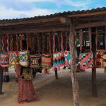 Del techo de un rústico puesto de madera cuelgan bolsas con estampados de colores, entre las que se encuentra una persona con falda larga. El suelo es arenoso y el cielo está parcialmente nublado.