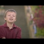 Un joven con un polo granate sonríe con los ojos parcialmente cerrados. Detrás de él, la vegetación borrosa y las escaleras apuntan a la cercana Biblioteca Pública Municipal Isaías Naranjo.