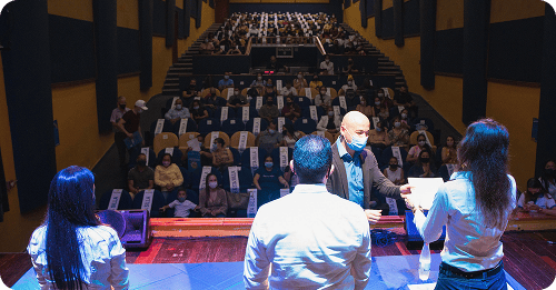 La gente está reunida en el Teatro Comfamiliar. Algunos individuos están en el escenario frente al público, sentado y separado, y una persona enmascarada recibe un documento de otra.