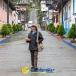 Una persona mayor con sombrero y chaqueta está de pie en la tranquila y colorida calle de La Celia sosteniendo un paquete rosa, rodeada de árboles recortados; el logotipo de Comfamiliar Risaralda aparece en la parte inferior, cerca de la señalización de la Biblioteca Pública.