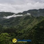 Exuberantes montañas verdes bajo un cielo nublado con niebla flotando por los valles cercanos a La Celia; el logotipo y el texto Comfamiliar aparecen en la parte inferior de la imagen.