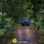 Un todoterreno azul circula por un estrecho camino de tierra rodeado de un denso follaje verde selvático, creando un ambiente aventurero y tropical. Los logotipos de Confamiliar y Comfamiliar La Celia aparecen en la parte inferior.