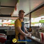 Un hombre con camisa amarilla, delantal y gorra detrás del mostrador de un bar de zumos al aire libre en La Celia, rodeado de batidos recién hechos. El logotipo de la Biblioteca Pública Comfamiliar aparece en la parte inferior de la imagen.