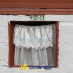 Una pequeña ventana con cortinas blancas de encaje en una pared blanca con marco marrón. Encima de la ventana hay un letrero verde con la dirección 2-24. En la parte inferior está el logotipo de Comfamiliar Risaralda, que destaca su conexión con la Biblioteca Pública La Celia.