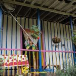 Una persona está de pie en un colorido porche con paredes de rayas vibrantes, plantas en macetas y un banco, utilizando un palo largo para ajustar una maceta colgante. En la parte inferior se ve la marca familiar, que añade encanto a este escenario de La Celia.