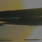 Vista al atardecer de una tranquila masa de agua con reflejos, siluetas de edificios y estructuras de madera a lo largo de la orilla, y un suave resplandor dorado en el cielo.