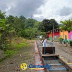 Una mujer conduce un pequeño carro sobre raíles a través de una zona rural con frondosos árboles y casas de colores. El cielo nublado asoma por encima, mientras que el logotipo de Confamiliar y la Biblioteca Pública Municipal León de Greiff son visibles en la parte inferior de la imagen.