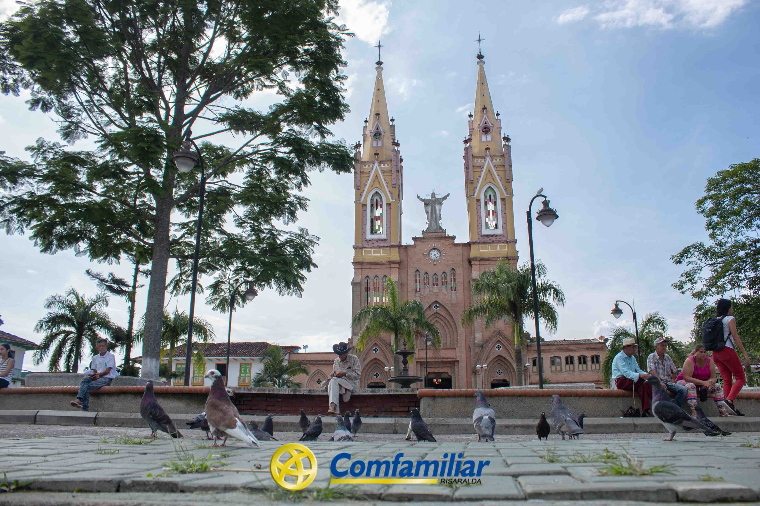 Una iglesia con dos altas torres da a una plaza cercana a la Biblioteca Pública Municipal, donde la gente se sienta en bancos y las palomas pasean en primer plano. Los árboles enmarcan la escena, y un logotipo azul de Comfamiliar Risaralda aparece en la parte inferior central.