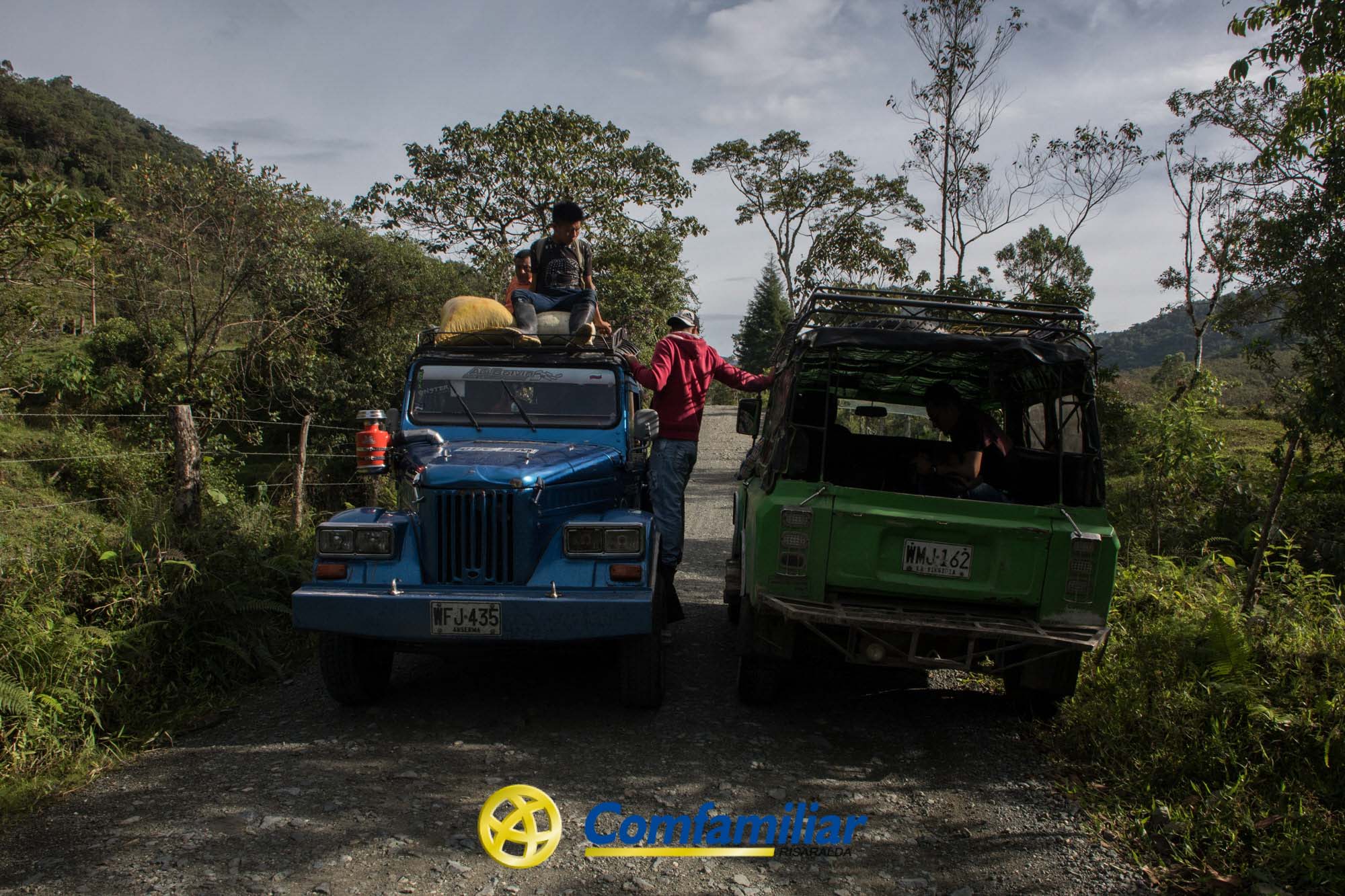 Dos jeeps se detienen en un camino rural de tierra rodeado de árboles y colinas en Mistrató; la gente se para y se sienta en el jeep azul, mientras que el jeep verde está aparcado a su lado. En la parte inferior aparece el logotipo de la Biblioteca Pública Comfamiliar.