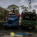 Dos jeeps se detienen uno junto al otro en una carretera rural de Mistrató rodeada de árboles. Tres personas están de pie o sentadas en el jeep azul, mientras que el jeep verde está ocupado. La luz del sol se filtra entre el follaje, añadiendo encanto a esta tranquila escena.