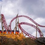 En el Parque del Café, una montaña rusa roja llamada Krater serpentea a través de bucles y giros bajo un cielo nublado, con su nombre escrito en negrita sobre una base rocosa pintada de amarillo en primer plano.