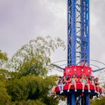 Un grupo de personas se sientan en una atracción roja del Parque del Café, preparándose para ascender a una alta torre azul, rodeados de árboles verdes bajo un cielo nublado.