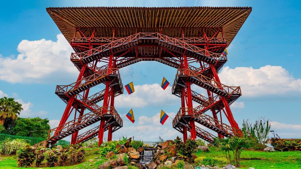 Gran torre de observación de madera con vigas y escaleras rojas, rematada por un amplio tejado plano. Banderas de Colombia cuelgan entre las escaleras del Parque del Café. Bajo un cielo azul, se puede ver una exuberante vegetación y una pequeña cascada.