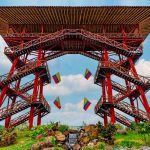 Gran torre de observación de madera con vigas y escaleras rojas, rematada por un amplio tejado plano. Banderas de Colombia cuelgan entre las escaleras del Parque del Café. Bajo un cielo azul, se puede ver una exuberante vegetación y una pequeña cascada.