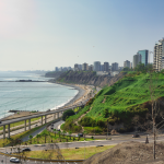 Un paisaje urbano costero con modernos rascacielos sobre verdes acantilados, carreteras curvas debajo y una playa de arena junto al océano azul bajo un cielo despejado.