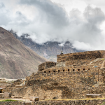 Unas antiguas ruinas de piedra se alzan sobre un telón de fondo de montañas neblinosas y cielo nublado, con una persona visible en lo alto del nivel más alto de la estructura.