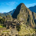 Las antiguas ruinas de piedra de Machu Picchu se asientan sobre una verde cresta montañosa, con terrazas y espectaculares picos elevándose al fondo bajo un cielo azul despejado.