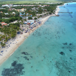 Vista aérea de una playa tropical con aguas turquesas, bañistas, palmeras y complejos turísticos cerca de la orilla. Muchas personas nadan y se relajan en la playa de arena bordeada de tumbonas.