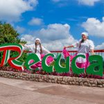 Dos mujeres con blusas blancas tradicionales y faldas rosas posan junto a un colorido cartel de Recuca, con vegetación y un cielo azul con nubes de fondo.