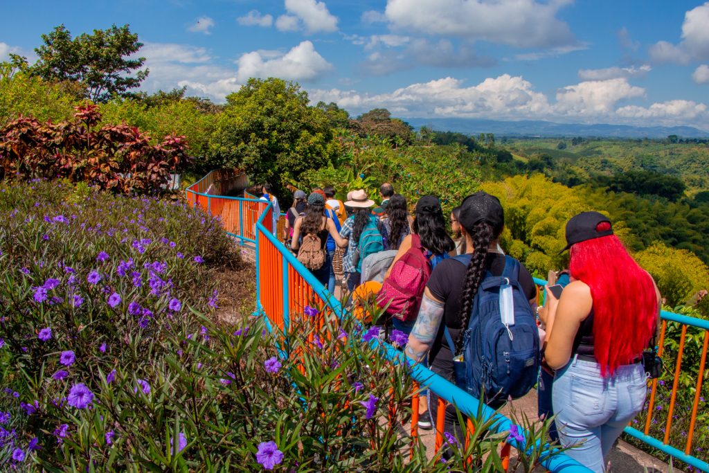 Un grupo de personas con mochilas camina por un sendero bordeado de flores moradas y barandillas de color azul anaranjado, rodeado de exuberante vegetación y con vistas a un pintoresco valle bajo un cielo parcialmente nublado.