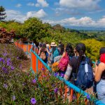 Un grupo de personas con mochilas camina por un sendero bordeado de flores moradas y barandillas de color azul anaranjado, rodeado de exuberante vegetación y con vistas a un pintoresco valle bajo un cielo parcialmente nublado.