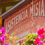 Un cartel rojo desgastado en el que se lee "Restaurante Asistencia Misia Gumercinda" queda parcialmente oculto por unas flores de color rosa intenso en primer plano.