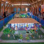 Un grupo de personas observa una colorida maqueta en miniatura de una casa y un jardín tradicionales, expuesta en el interior de un edificio de madera con puertas y ventanas de color rojo y azul brillante. En la maqueta hay árboles, animales y figuras.