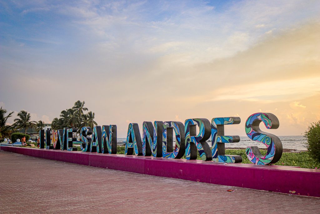 Grandes letras de colores deletrean VIVE SAN ANDRÉS a lo largo de un paseo marítimo al atardecer, con palmeras y el océano visibles al fondo.