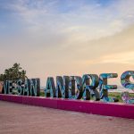 Grandes letras de colores deletrean VIVE SAN ANDRÉS a lo largo de un paseo marítimo al atardecer, con palmeras y el océano visibles al fondo.