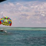 Un paravela de colores brillantes con la palabra Richie flota sobre una embarcación en aguas turquesas del océano bajo un cielo parcialmente nublado. Varias personas están en la barca y se ven arrecifes a lo lejos.
